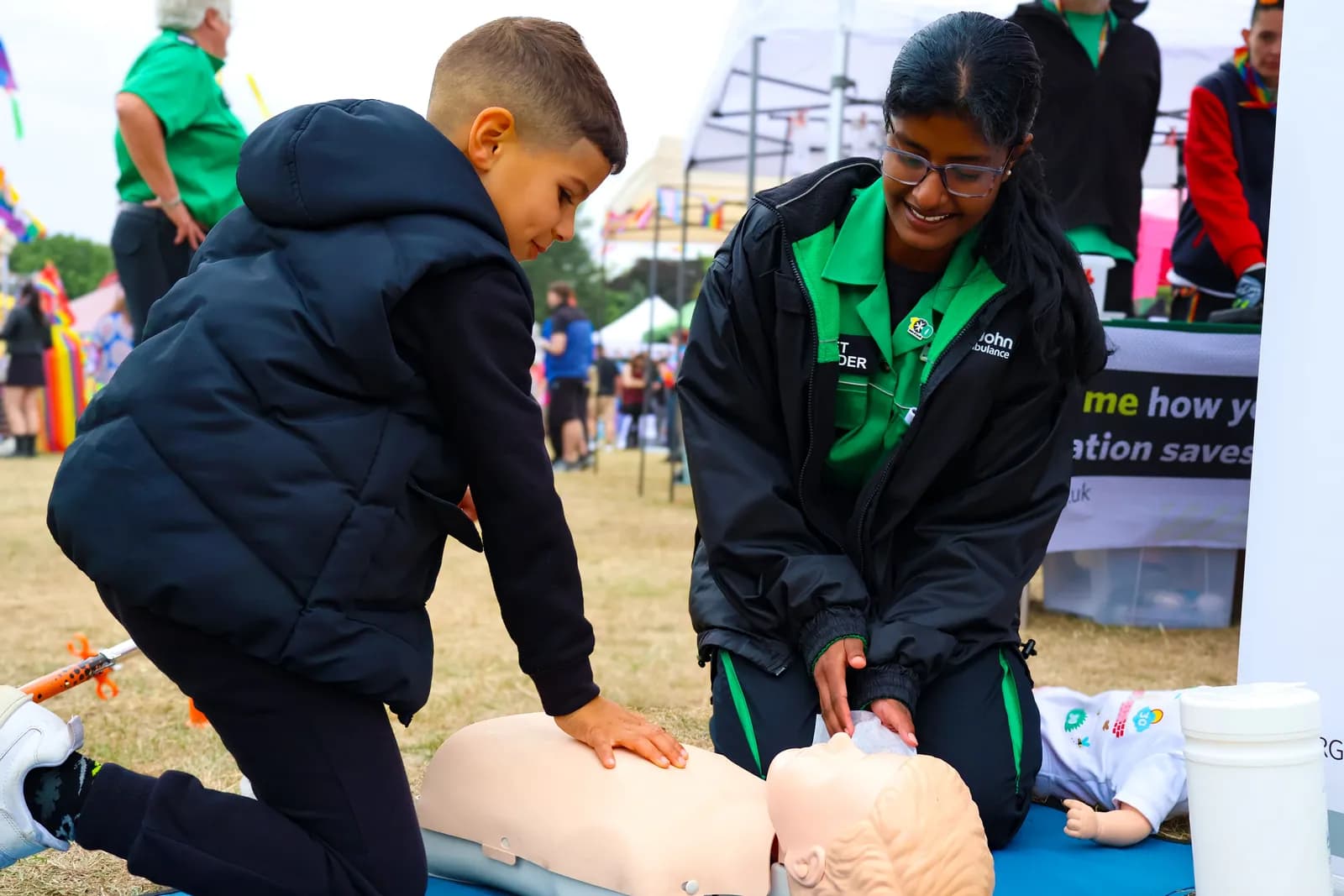 A young boy and a woman in a St John Ambulance uniform are practicing CPR on mannequins at an outdoor event.