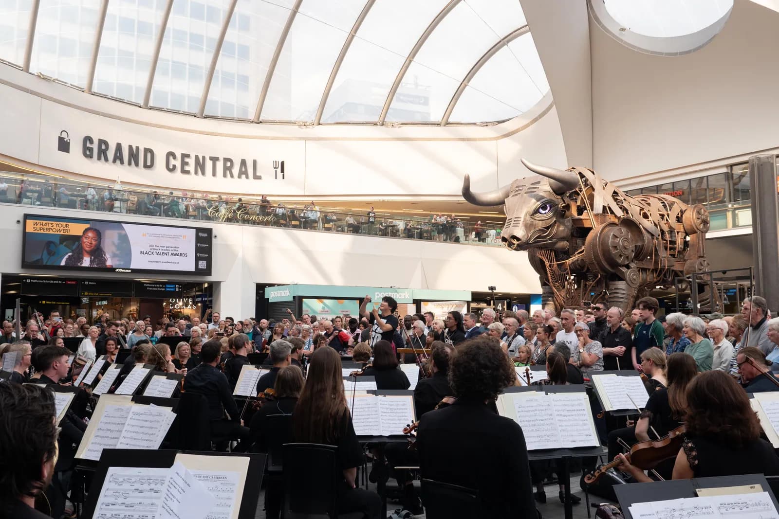 An orchestra performs in front of a crowd and a large mechanical bull statue inside Grand Central Station.