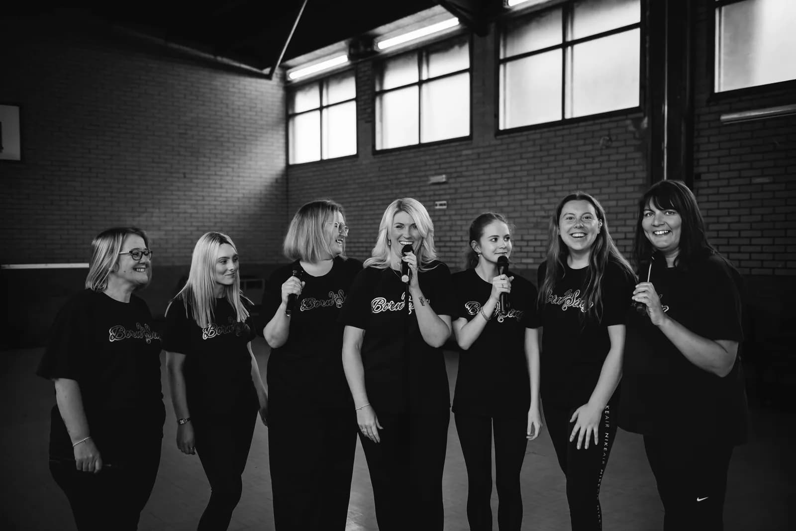 Seven women in black t-shirts and trousers hold microphones and sing or speak, with a brick wall and windows in the background.