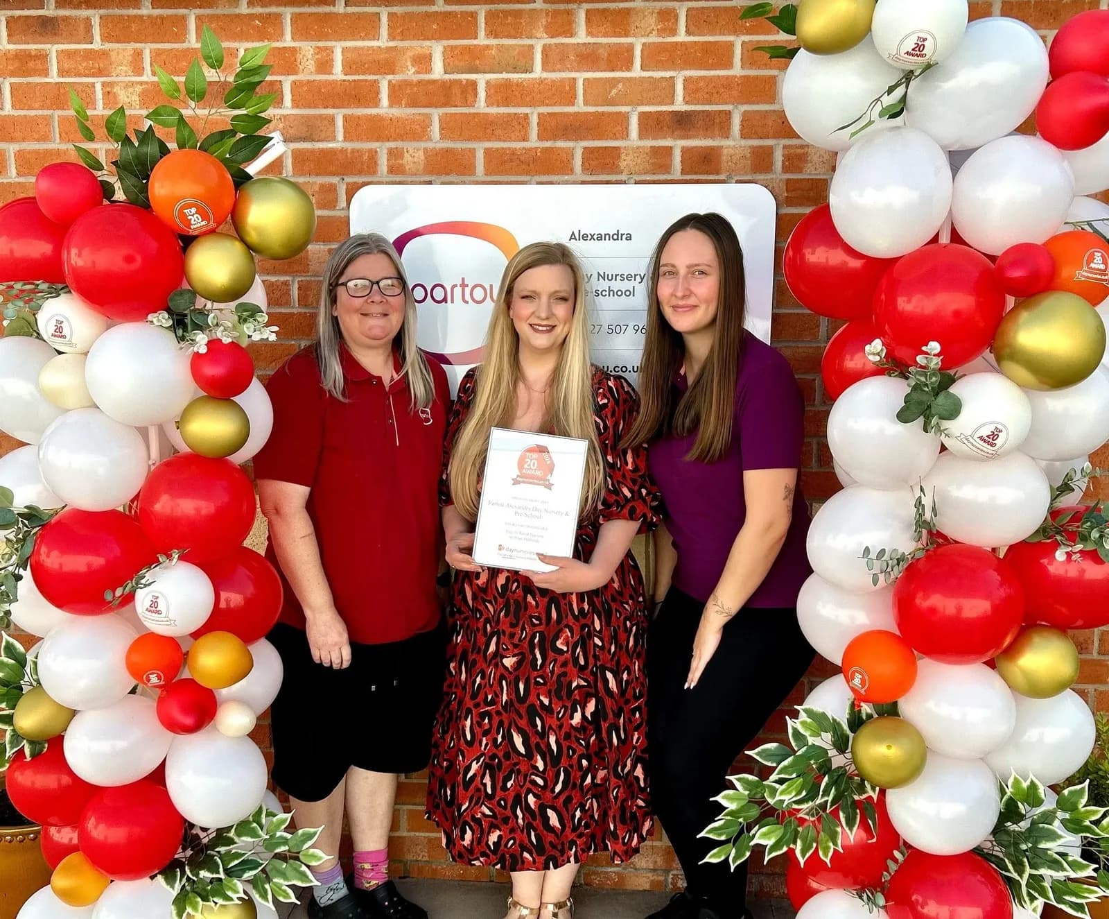 Three women stand smiling in front of a brick wall adorned with red, white, and gold balloons; the woman in the center holds a framed certificate.