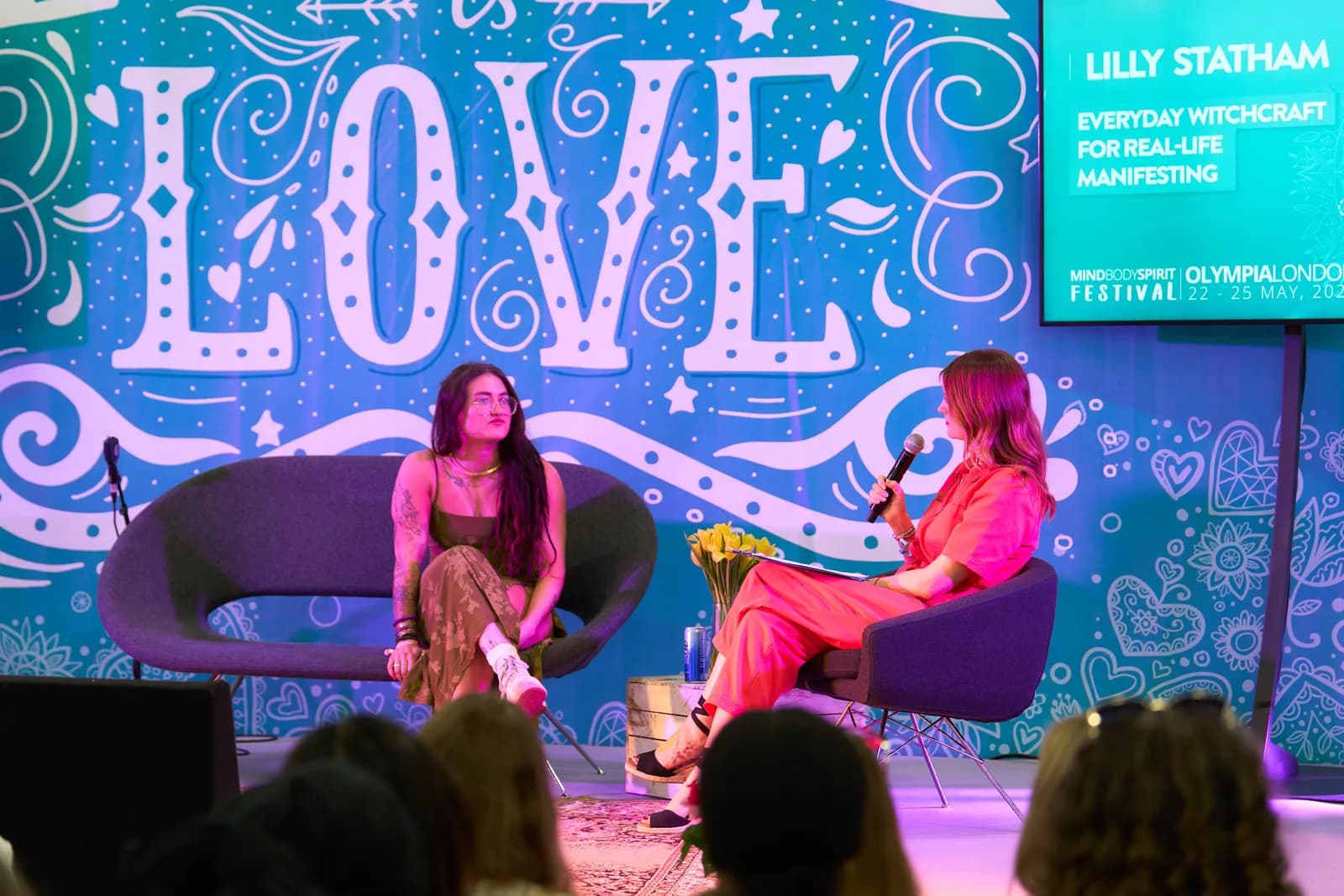 Two women, one with long dark hair and the other with shoulder-length reddish-brown hair, sit on chairs facing each other on a stage in front of a blue backdrop with white "LOVE" lettering and various designs. The woman on the right holds a microphone and appears to be interviewing the woman on the 