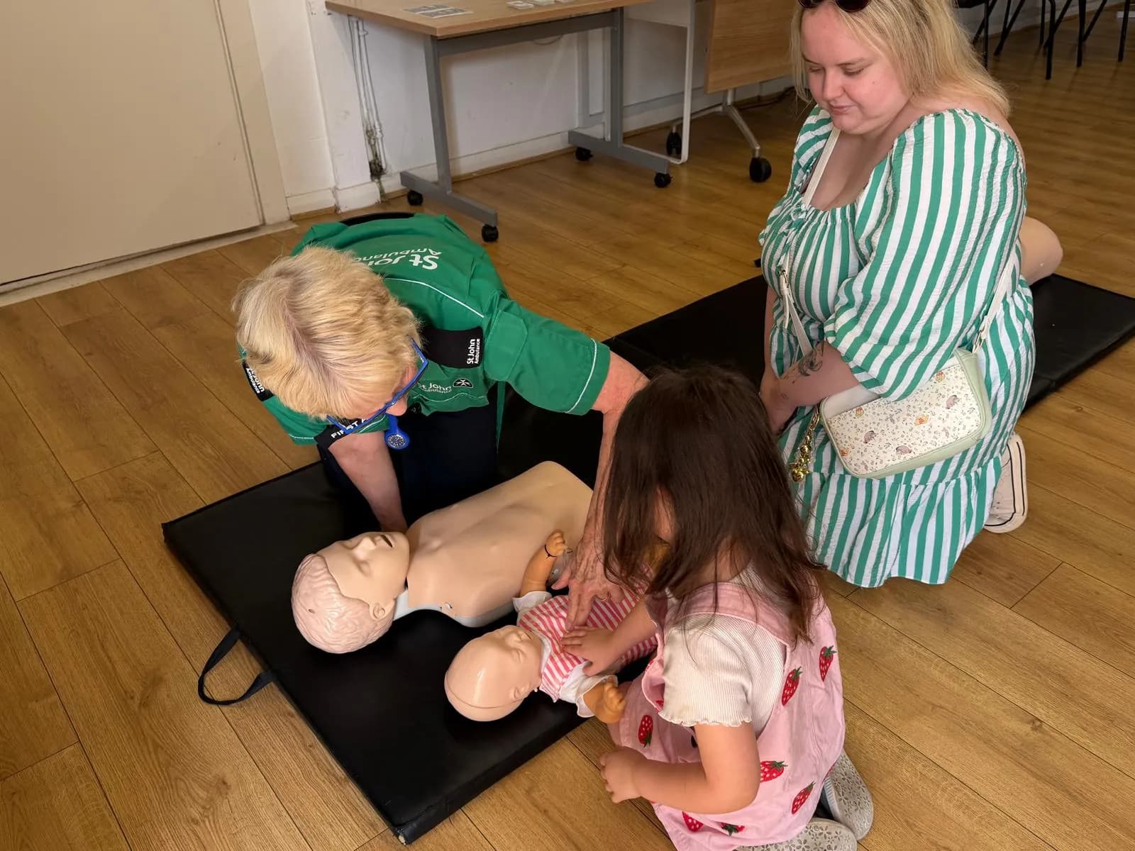 A woman in a green uniform demonstrates CPR on an adult mannequin while a young girl attends to a baby mannequin, with another woman observing.