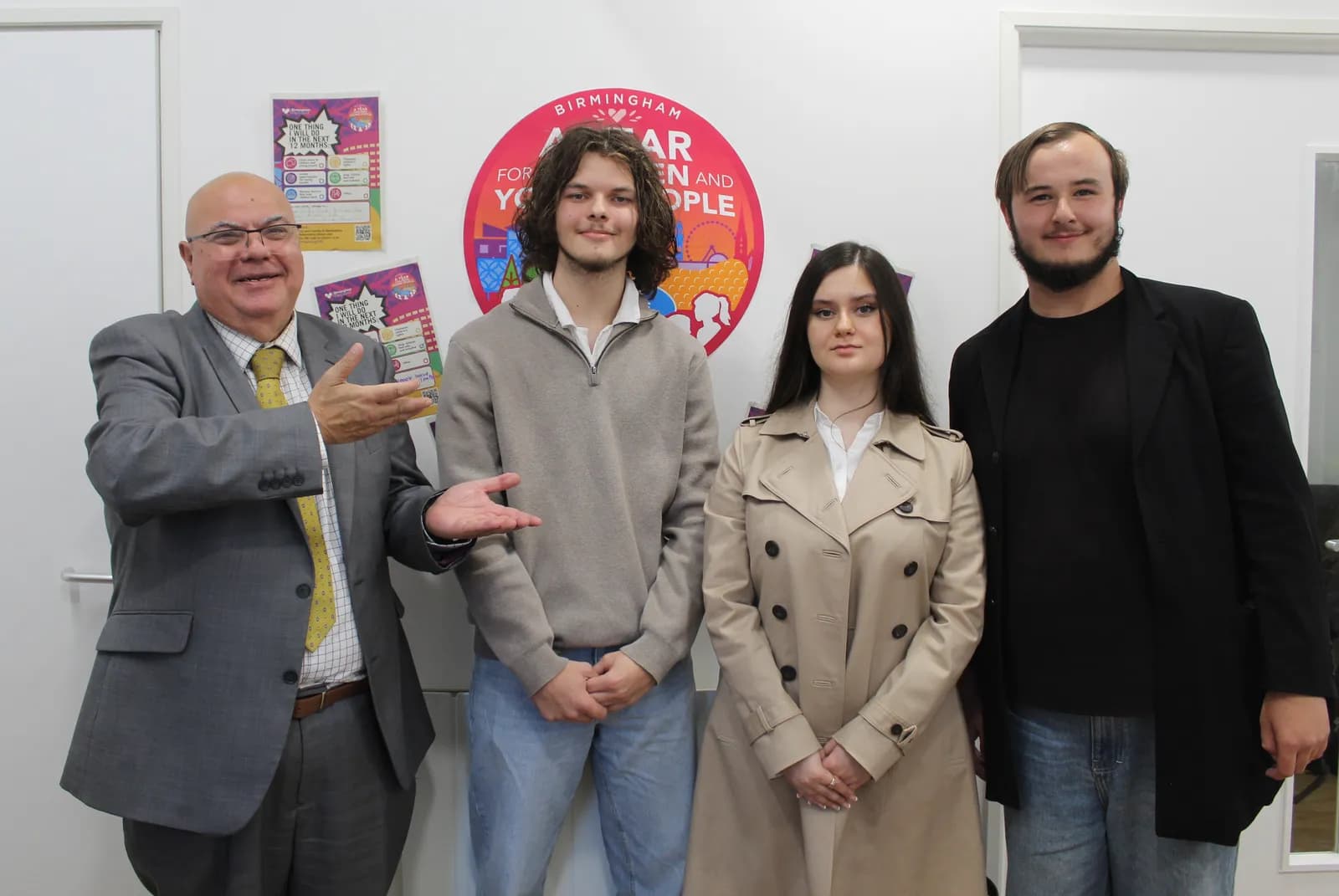 Four people, including an older man in a suit gesturing, stand in a line in front of a white wall with posters and a circular red logo.